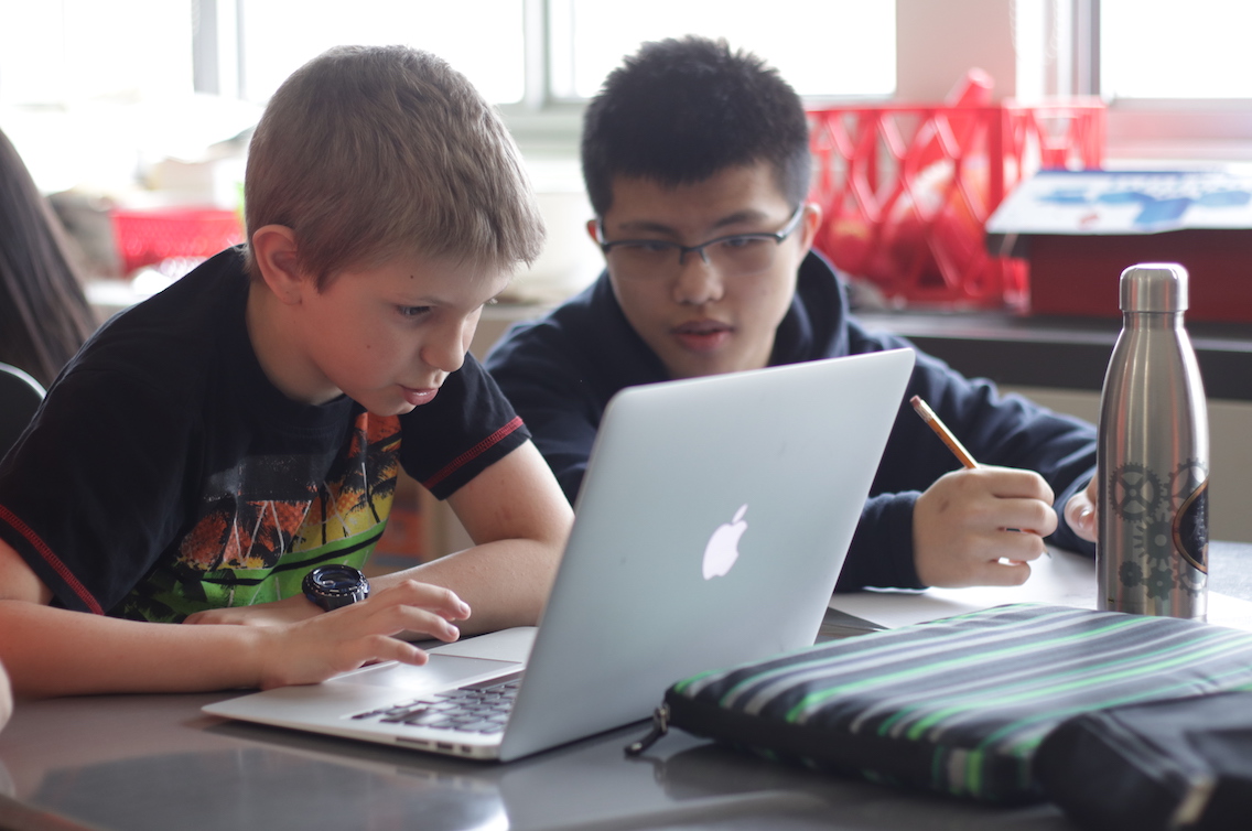 A grade four student operates a computer while a grade ten student looks on.