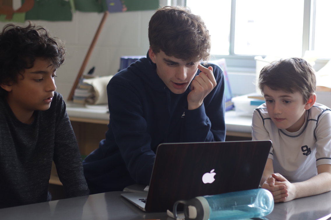 Two younger students listen while an older students explains some code running on a computer.